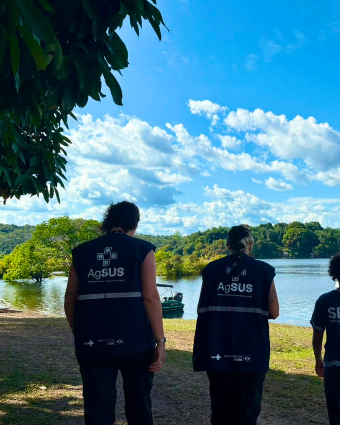 Três profissionais de colete da AgSUS diante de um rio, de costas, olhando o território